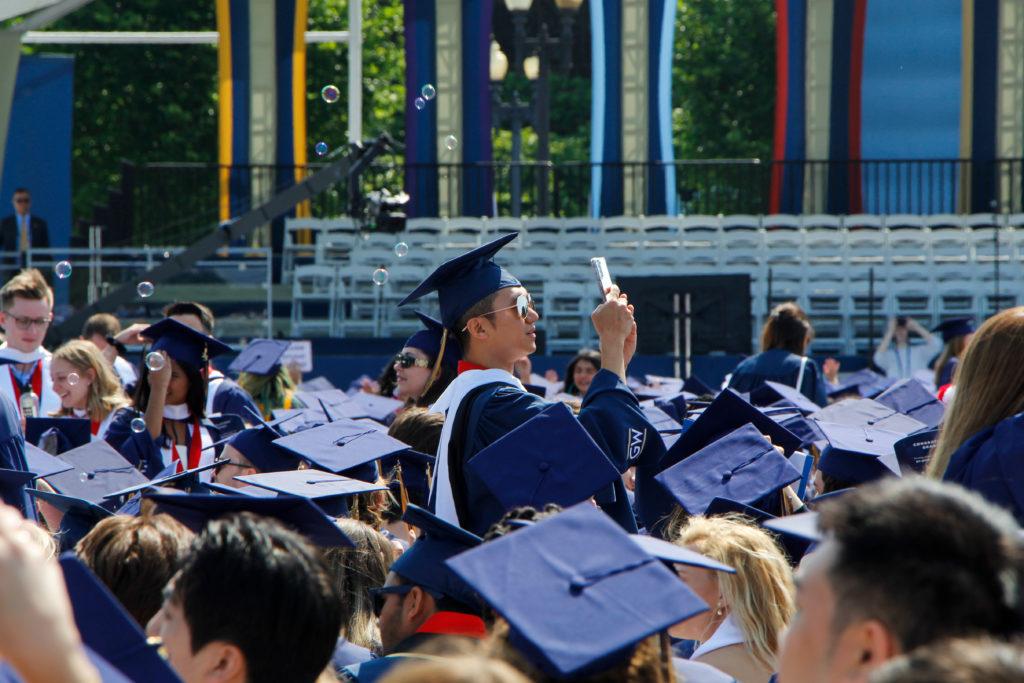 A graduate takes a photo of the Commencement scene while bubbles float over the crowd. 